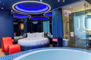 Een moderne hotelkamer met een rond bed, een douchecabine van glas en een badkuip met blauwe tegels.
