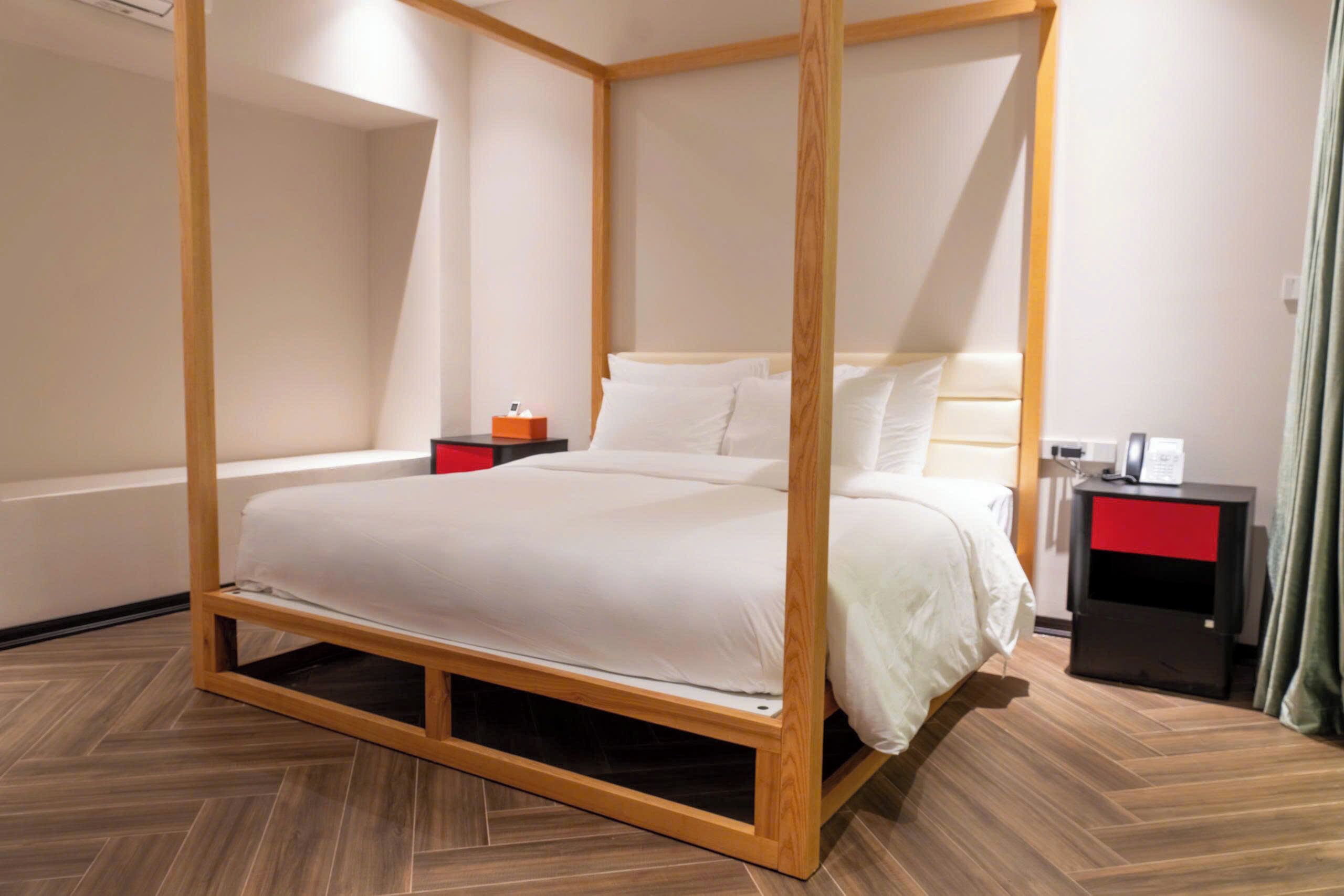 Un lit à baldaquin avec une structure en bois et une literie blanche, une table de chevet avec un tiroir rouge et une petite table de nuit avec un téléphone et une lampe.