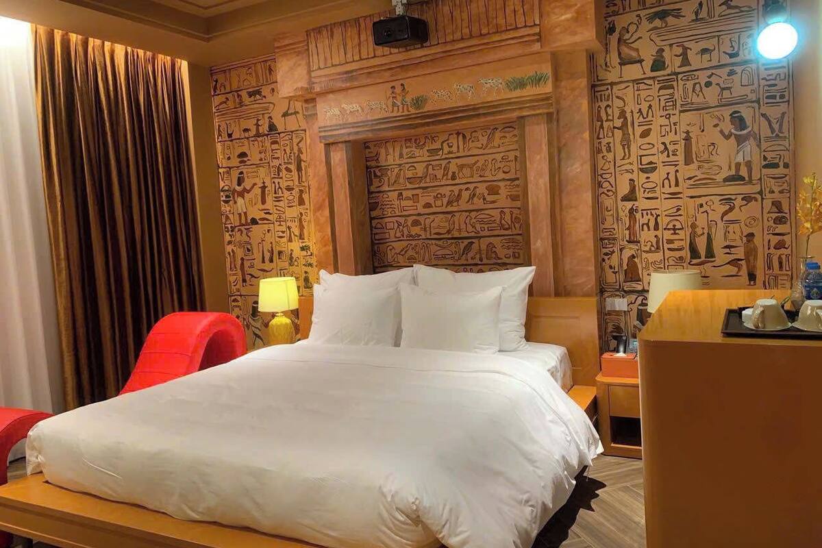 A hotel room with a large bed, a red chair, a wooden headboard with hieroglyphs, a nightstand with a lamp, and a desk with a cup and saucer.
