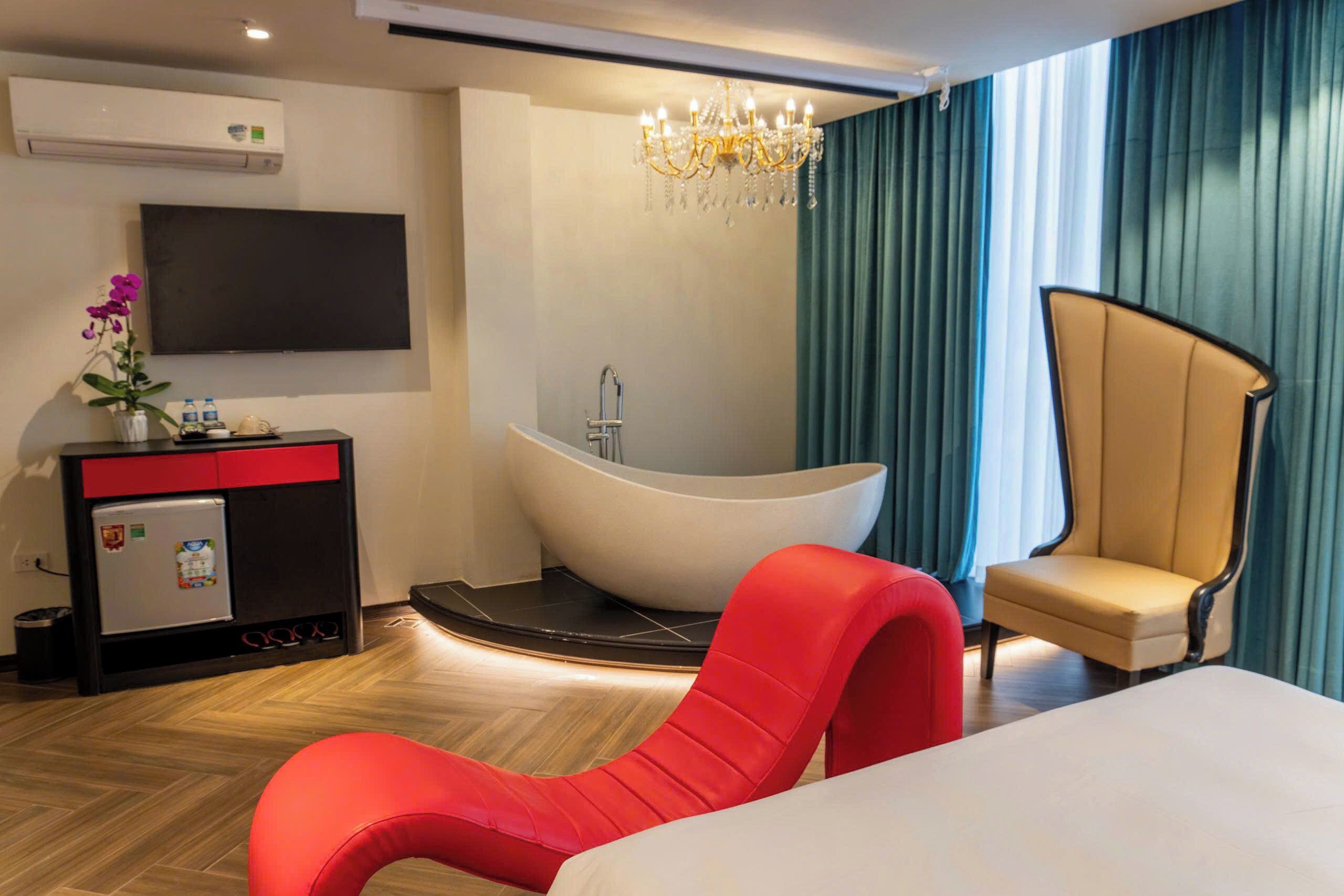 Una habitación de hotel moderna con una cama grande, un sillón rojo, una bañera, un televisor de pantalla plana y un minibar.