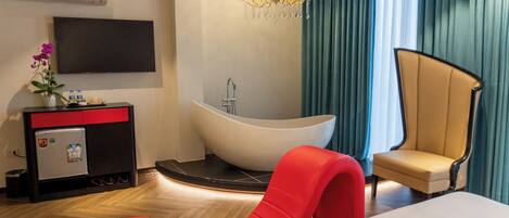 Una habitación de hotel moderna con una cama grande, un sillón rojo, una bañera, un televisor de pantalla plana y un minibar.