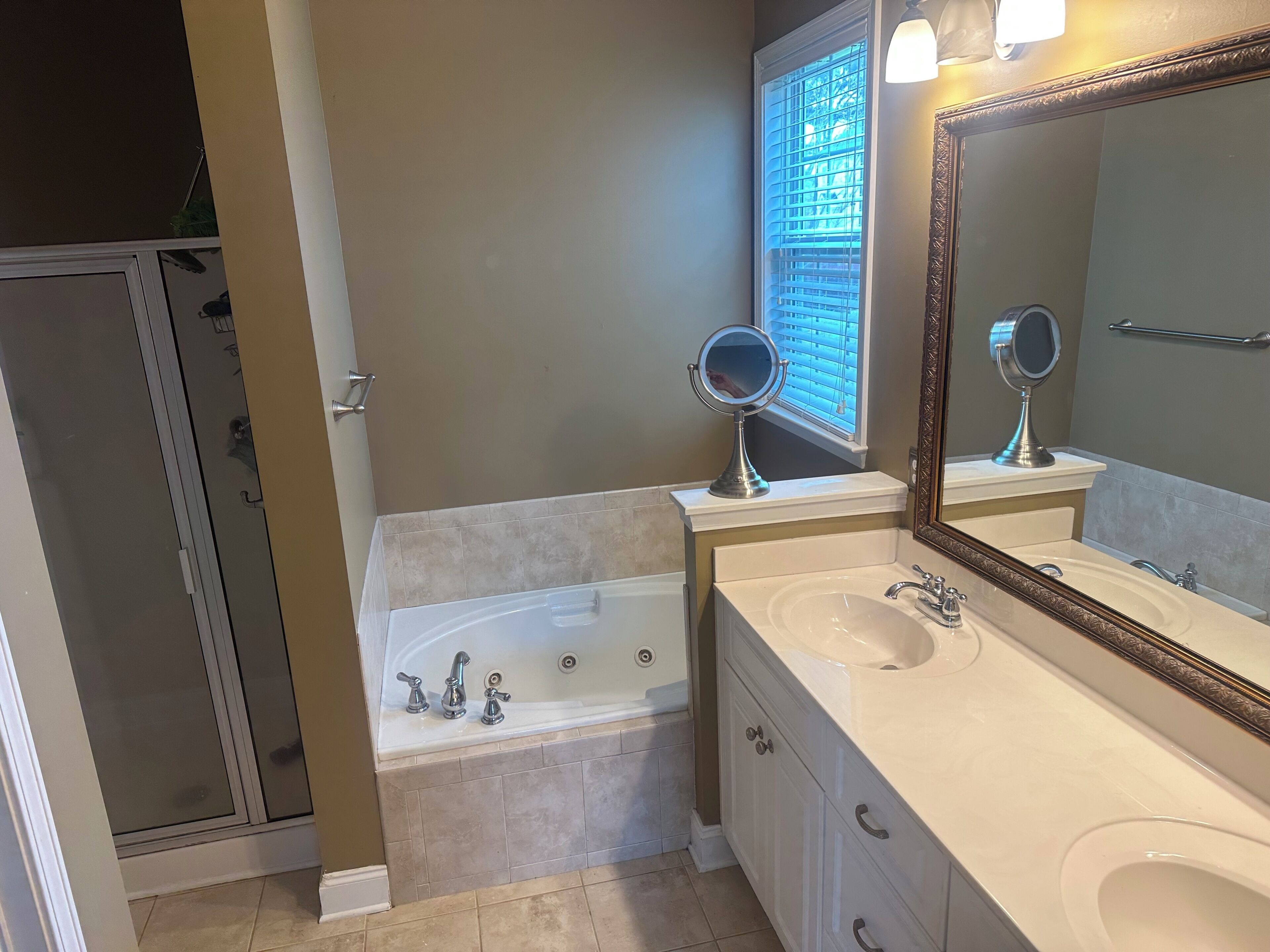 Une salle de bain avec une baignoire, un lavabo, deux miroirs et une fenêtre avec des stores.