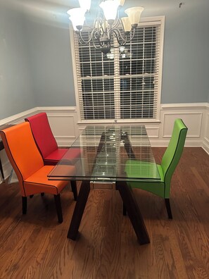 Een eetgedeelte met een tafel met glazen blad, twee stoelen met verschillende bekleding, een raam met jaloezieën en een kroonluchter.