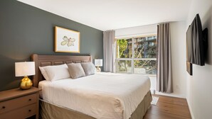 Un dormitorio con cama, mesitas de noche, lámpara, un cuadro enmarcado, ventana con cortinas y televisor montado en la pared.