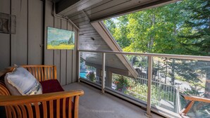 Una terraza de madera con barandilla de vidrio, un cuadro en la pared y vistas a árboles y una piscina.