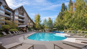 Una piscina con sillones de descanso, una zona de estar cubierta y un edificio con balcones.