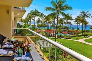 Un balcon aménagé avec un coin repas, donnant sur une piscine entourée de palmiers et offrant une vue sur l’océan.