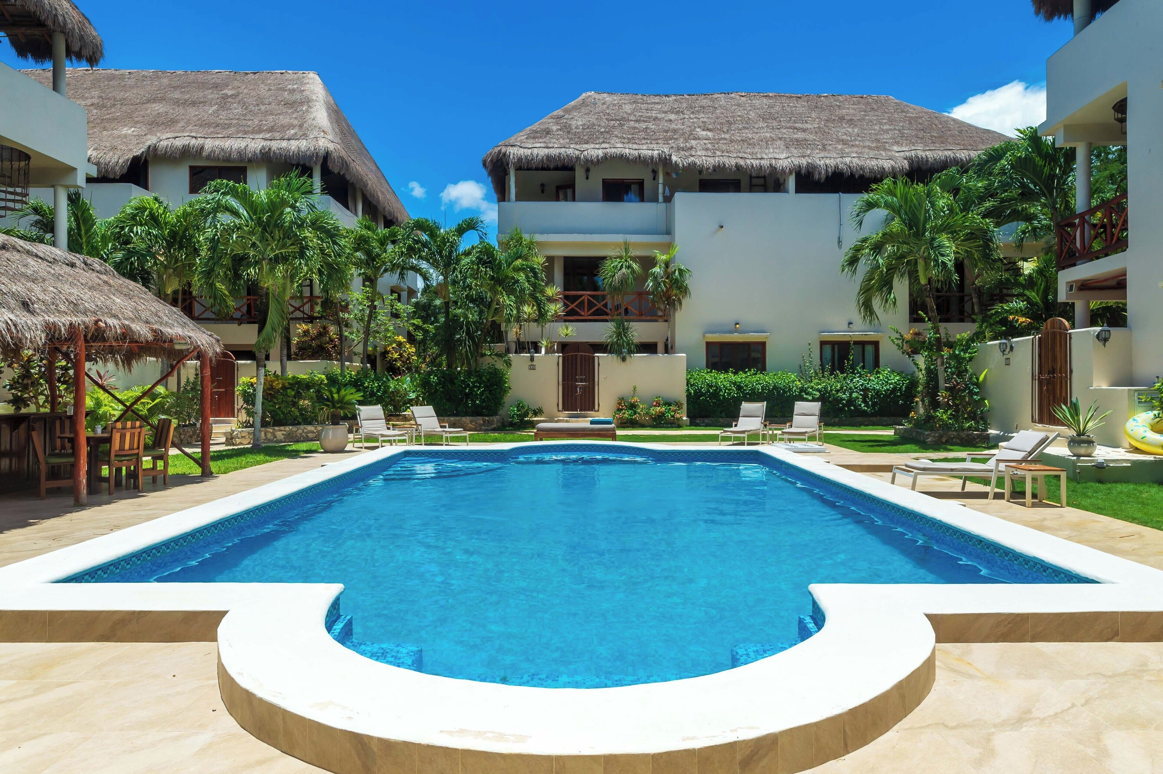 A swimming pool with lounge chairs, a thatched-roof building, and lush greenery.