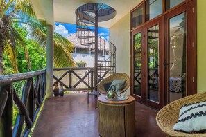 Varanda com escadaria em espiral, mobiliário de vime e vista para uma casa com telhado de palha.