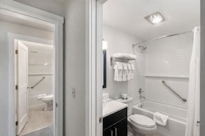Uma casa de banho moderna com chuveiro, sanita e lavatório. Existem barras de apoio no chuveiro e um porta-toalhas sobre o lavatório.