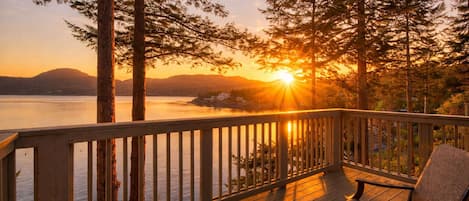 Una terraza de madera con barandilla, con vista a un lago y una puesta de sol al fondo.