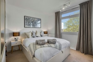 Ein Schlafzimmer mit einem großen Bett, Nachttischlampen, einem gerahmten Kunstwerk und einem Fenster mit Vorhängen.
