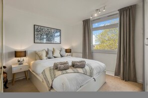 Ein Schlafzimmer mit einem Bett, Nachttischlampen, einem Fenster mit Vorhängen und einem gerahmten Bild an der Wand.
