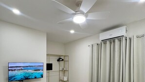 Hotellihuone, jossa on sänky, kattotuuletin, ilmastointilaite, televisio, seinälle kiinnitetty hylly, vaatekaappi ja verho.