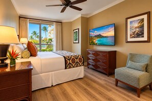 Habitación de hotel con cama, escritorio, silla, televisor y ventana con vista a palmeras y una playa.