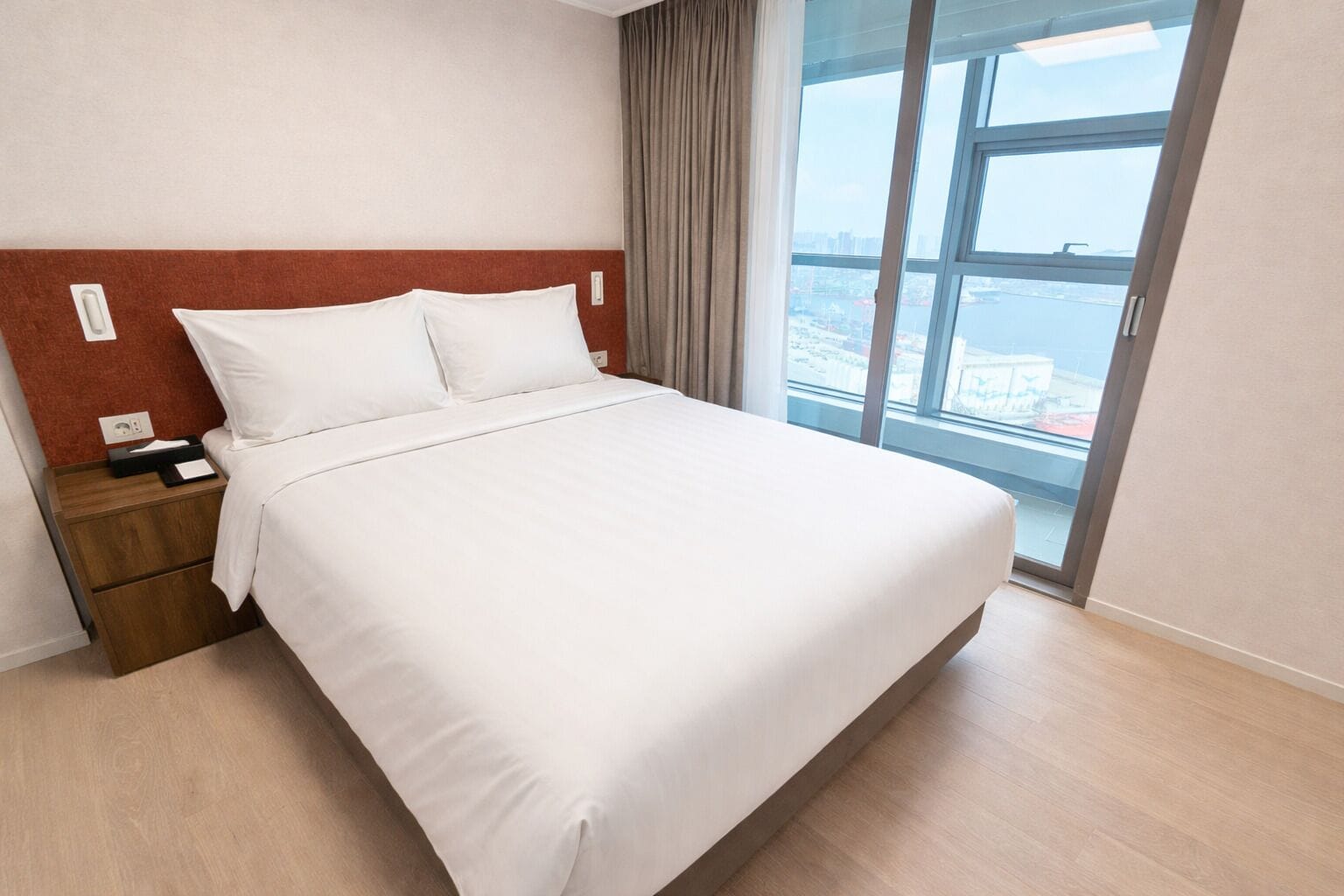 Quarto de hotel com uma cama grande, uma mesa de cabeceira, uma janela com vista para a cidade e uma parede de destaque vermelha.