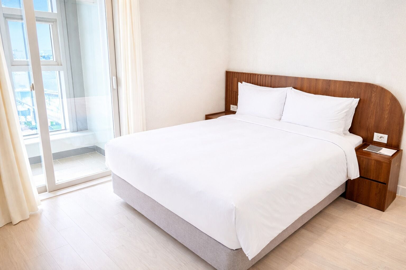 A hotel room with a large bed, wooden headboard, white bedding, a nightstand with a phone and a book, and a view through a glass door.