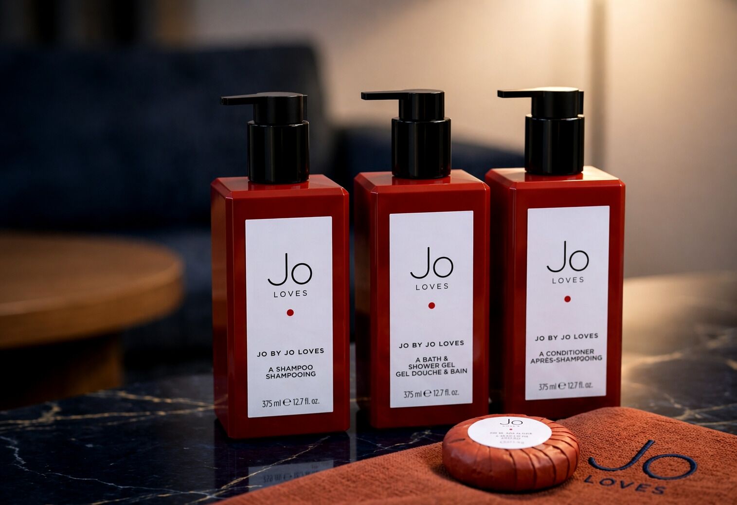 Three red bottles of Jo Loves shampoo and shower gel, a conditioner, and a bar of soap on a marble surface.