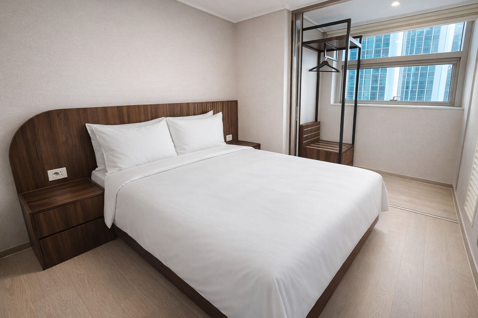 A modern hotel room with a wooden headboard, a large bed, a nightstand, a built-in wardrobe, and a view of the city through a large window.