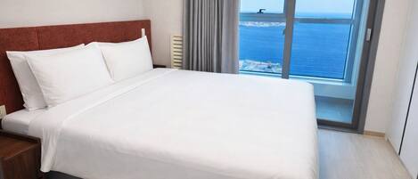 A hotel room with a bed, a nightstand, a window with a view of the sea, and a door.