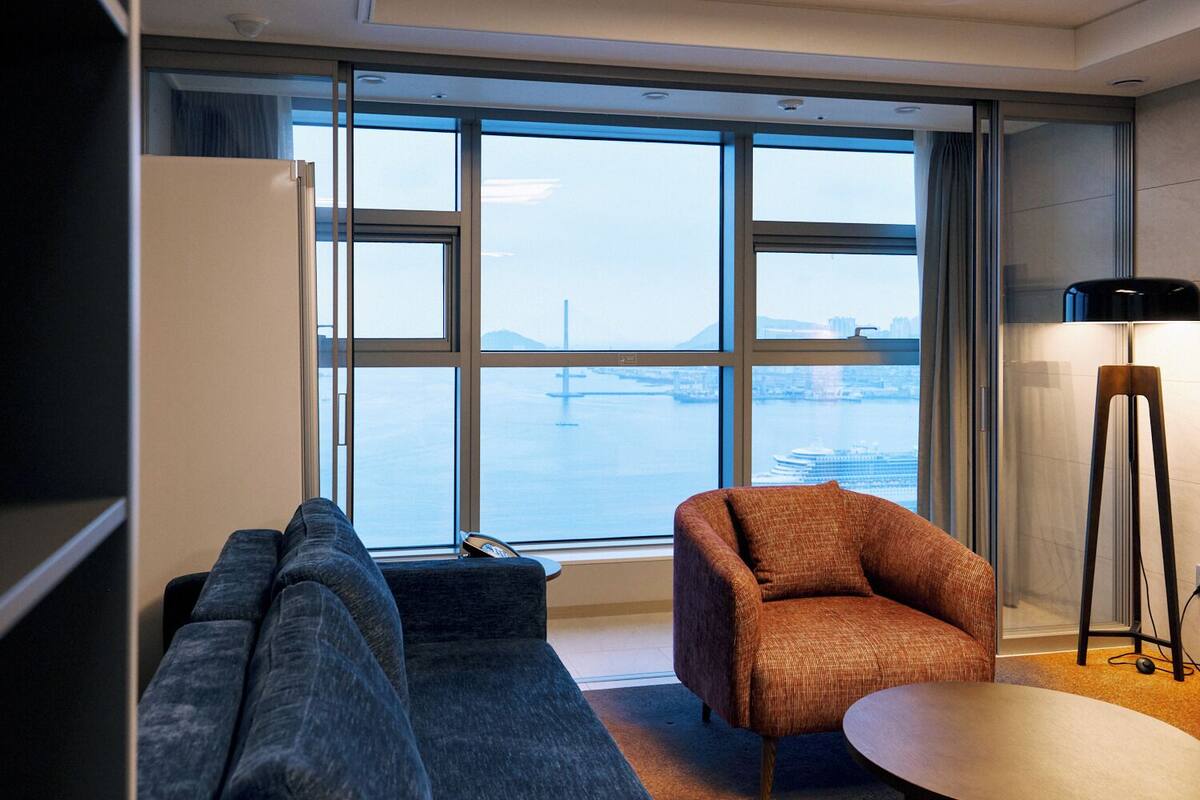 A modern room with a sofa, armchair, and floor lamp, large windows with a city view, and a small table.