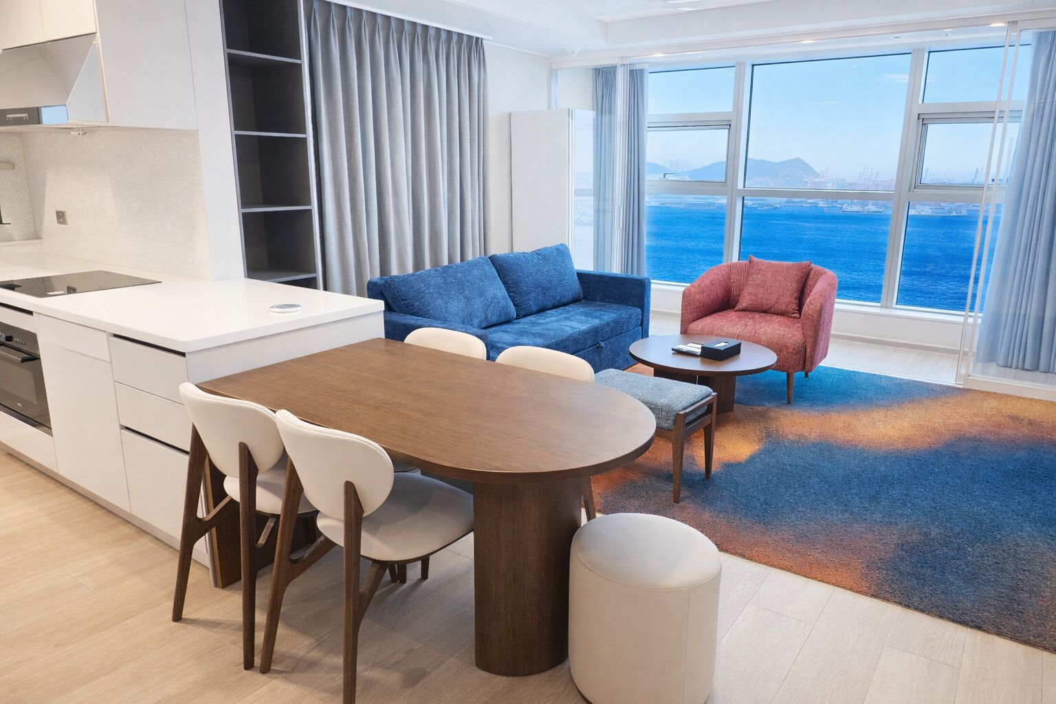 A modern living room with a dining table, two sofas, a kitchen area, and a large window with a view of the sea.