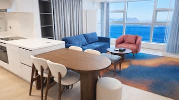 A modern living room with a dining table, two sofas, a kitchen area, and a large window with a view of the sea.