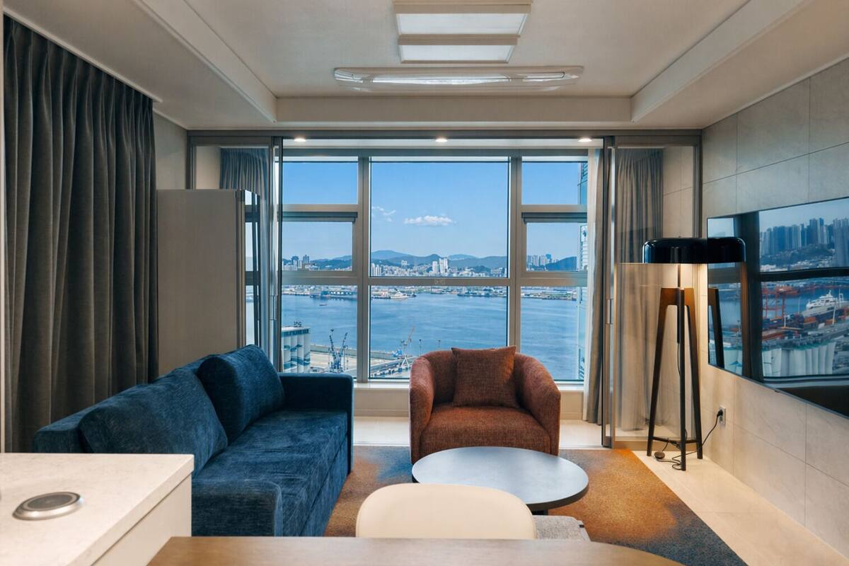 A modern hotel room with a large window, a sofa, a chair, a coffee table, and a flat-screen TV.