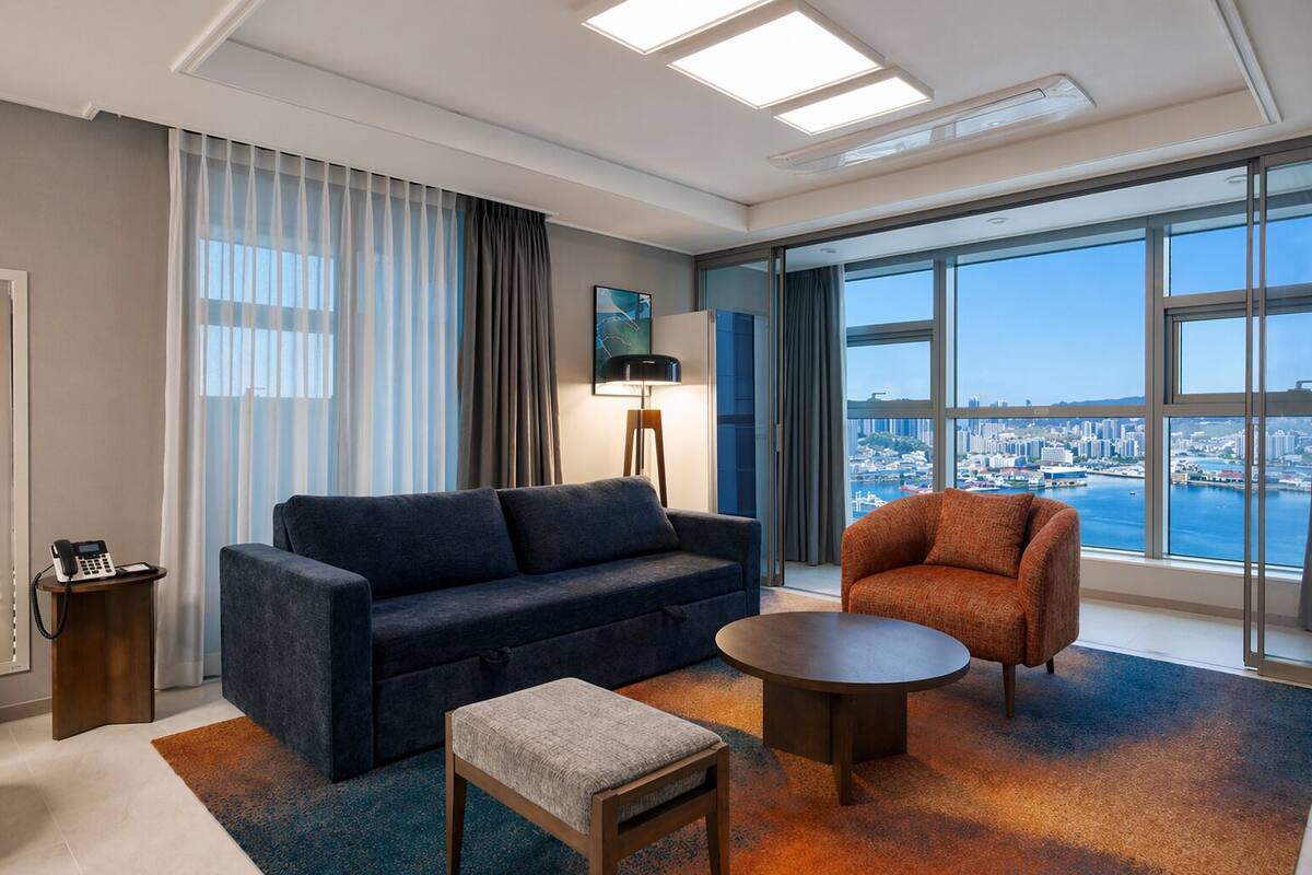 A hotel room with a sofa, armchair, small table, and a city view through large windows.