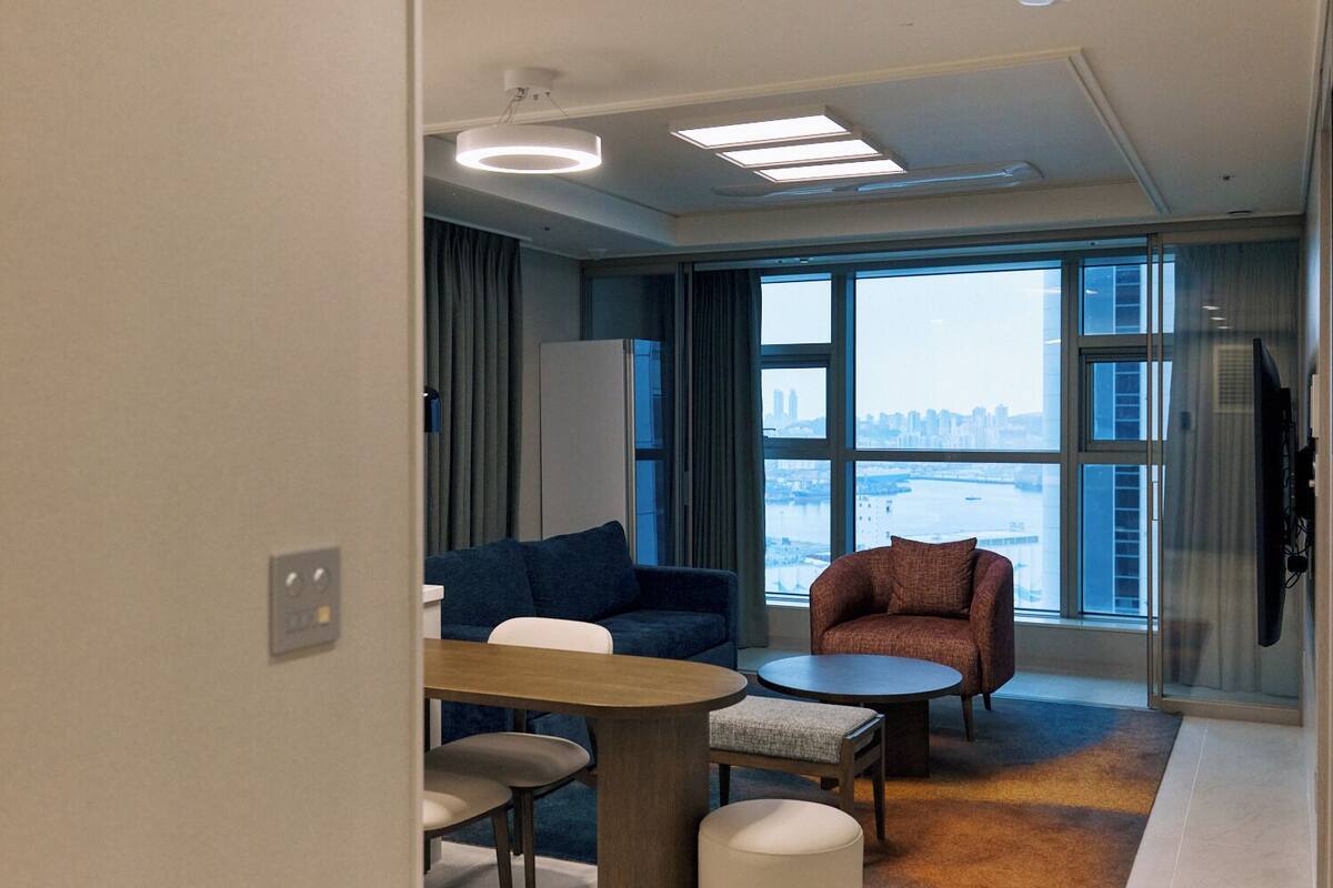A modern hotel room with a large window, a sofa, a chair, a small table, and a flat-screen TV.