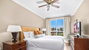 Habitación de hotel con cama, mesitas de noche, televisor, ventilador de techo y ventana con vista a palmeras.