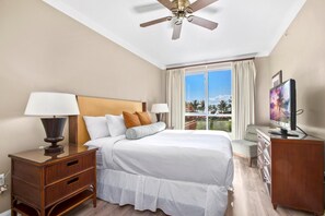 A hotel room with a bed, bedside tables, a TV, a ceiling fan, and a window with a view of palm trees. - Honua Kai | Ocean View 7 BR, Sleeps 24 | Car Incl. w/ 6+ Nights | HKK ML-3269 by KBM (Lahaina)