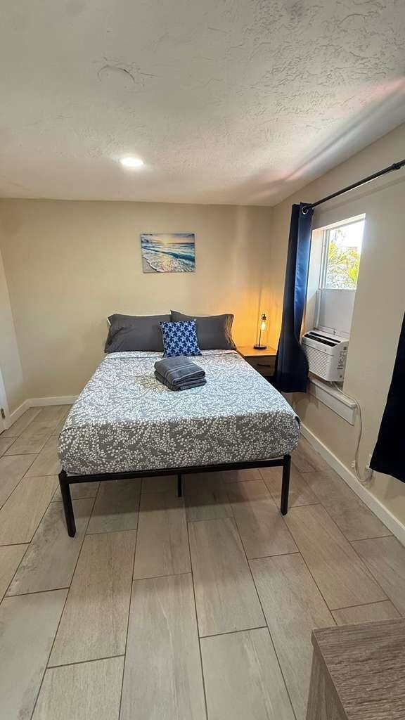 A bedroom with a bed, a nightstand, a lamp, a window with curtains, and a painting on the wall.