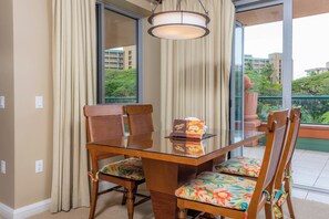 Habitación de hotel con una mesa de vidrio, sillas de madera, un cojín floral, una caja con temática hawaiana y vistas a zonas verdes y edificios.
