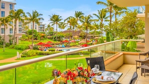 A balcony with a table set for dining, overlooking a pool area with palm trees and a lush garden. - Honua Kai | Ocean View 6 BR, Sleeps 20 | Car Incl. w/ 6+ Nights | HKK ML-3278 by KBM (Lahaina)
