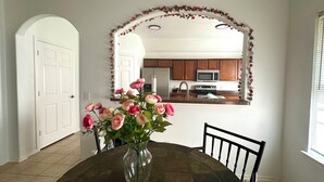 Un coin repas avec une table, des chaises, un vase de fleurs, une cuisine en arrière-plan et une porte blanche.