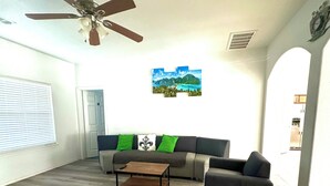 Un salon doté d’un ventilateur de plafond, d’un canapé avec des coussins verts, d’une table basse et d’où l’on aperçoit une cuisine à travers une arche.