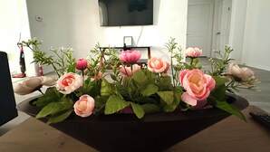 Un salon avec un téléviseur à écran plat fixé au mur, une plante en pot ornée de fleurs roses et une porte portant le numéro 3.