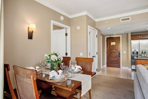 A dining area with a table set for four, a vase of flowers, and a kitchen visible in the background. - 2 Honua Kai Villas | Ocean View | Sleeps 10 | Free Car w/ 6+ Nights | ML-3281 (Lahaina)