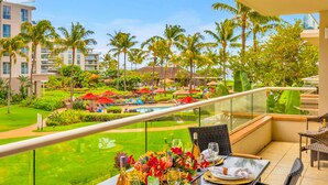 A balcony with a table set for dining, overlooking a pool area with palm trees and a lush garden. - Honua Kai | Ocean View 7 BR, Sleeps 24 | Car Incl. w/ 6+ Nights | HKK ML-3287 by KBM (Lahaina)