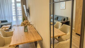 A modern living room with a wooden dining table, beige chairs, a sofa, and a large window with curtains. - A2 Modern 1-bed Apartment 2 (Plovdiv)
