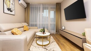 A modern living room with a sofa, a round coffee table, a TV mounted on the wall, and a large window with sheer curtains. - A2 Modern 1-bed Apartment 1 (Plovdiv)