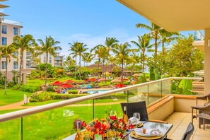 A balcony with a table set for dining, overlooking a pool area with palm trees and a lush garden. - Honua Kai | Ocean View 6 BR, Sleeps 20 | Car Incl. w/ 6+ Nights | HKK ML-3308 by KBM (Lahaina)