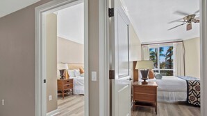A hotel room with a bed, bedside table, lamp, and a view of palm trees through the window. - Honua Kai | Ocean View 4 BR, Sleeps 14 | Car Incl. w/ 6+ Nights | HKK ML-3311 by KBM (Lahaina)