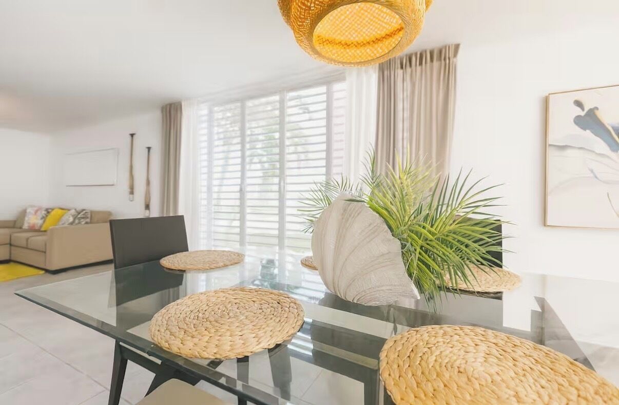A modern living room with a glass dining table, woven placemats, a large vase with green plants, a sofa, and a framed artwork on the wall.