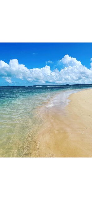 Ein Strand mit klarem Wasser, einem sandigen Ufer und einer entfernten Küste unter blauem Himmel mit Wolken.