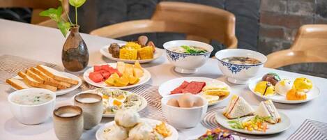 A table with various dishes, including bread, soup, and salads, set in a dining area with chairs and a brick wall in the background.