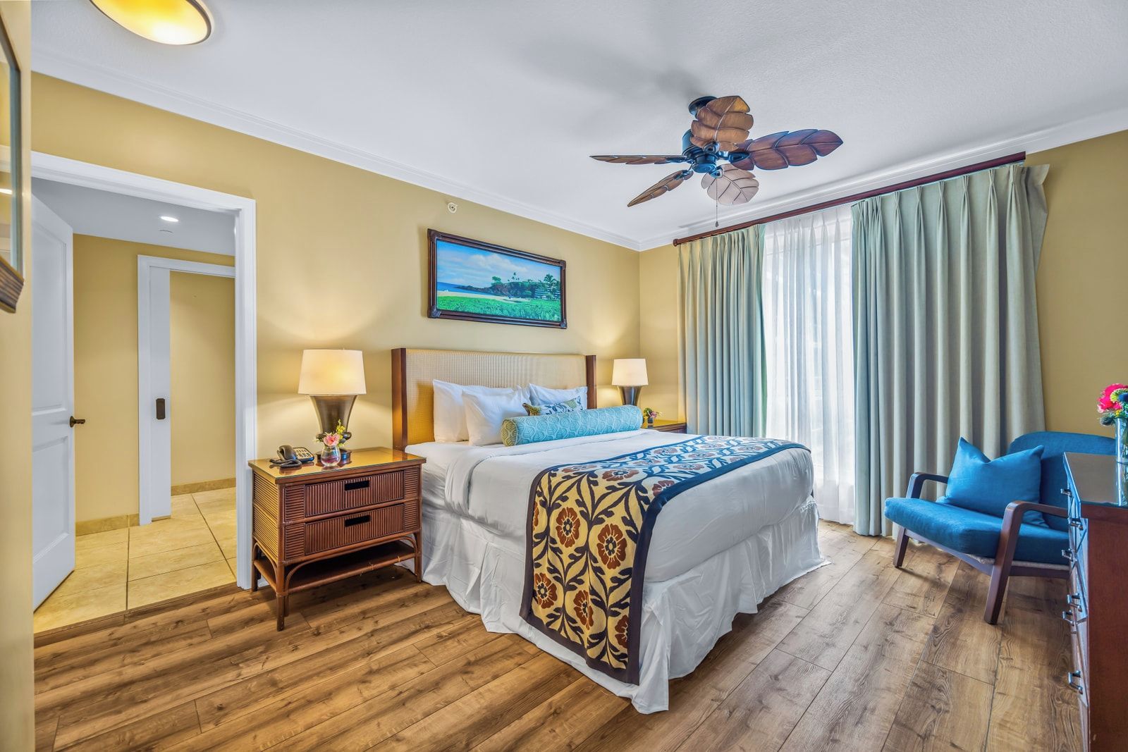 A hotel room with a bed, a desk, a chair, a ceiling fan, and a painting on the wall.