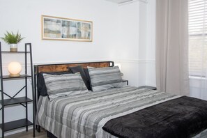 A neatly arranged bedroom with a bed, a bedside lamp, a framed artwork on the wall, and a potted plant.