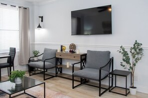 A modern living room with a flat-screen TV, two grey armchairs, a wooden side table, a small plant, and a glass coffee table.